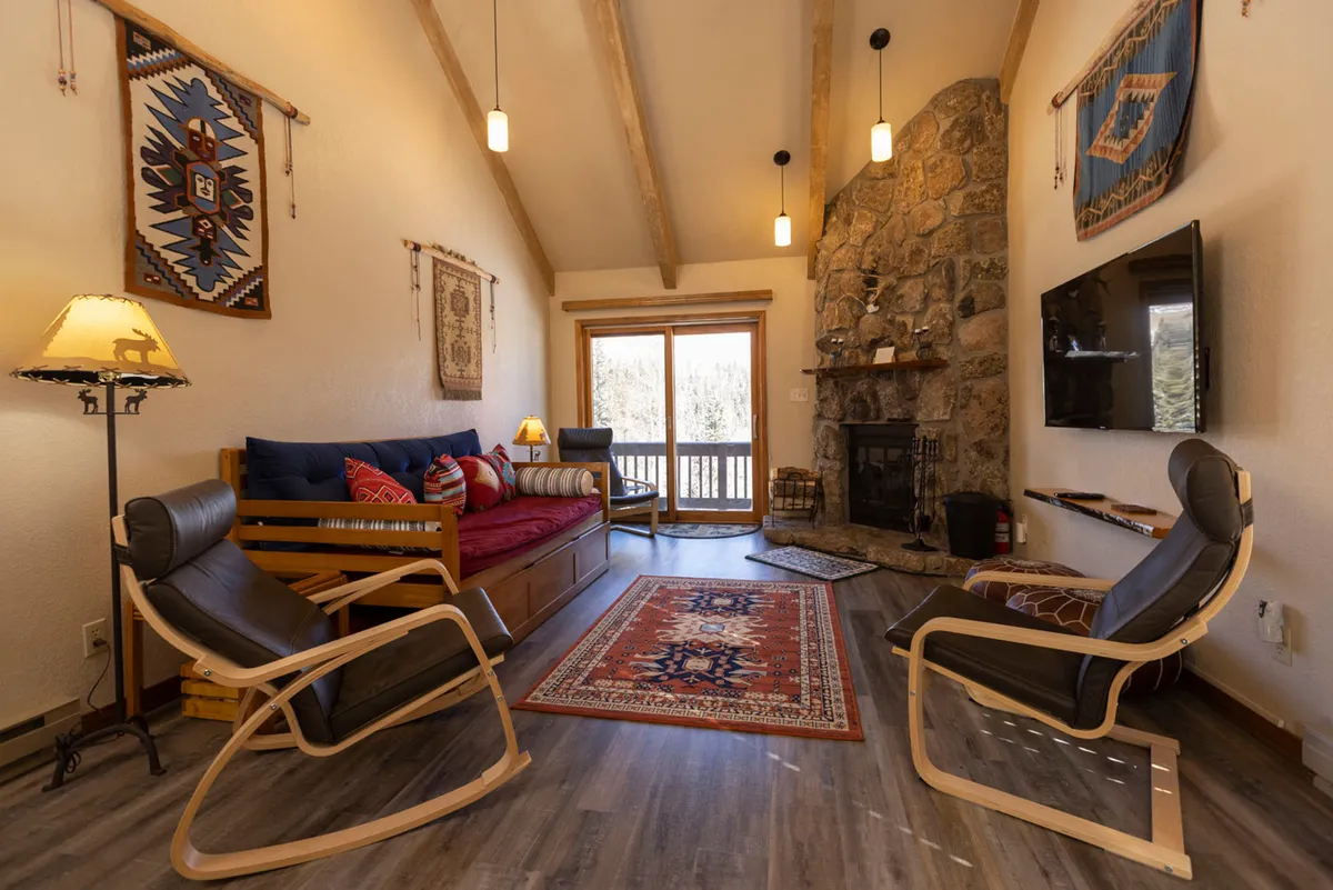 A welcoming seating area centered around a stone fireplace, accented by a decorative rug and wooden furnishings. Soft natural light pours in through the large windows, offering a serene spot for reading, relaxing, or enjoying quiet moments after a day on the slopes.