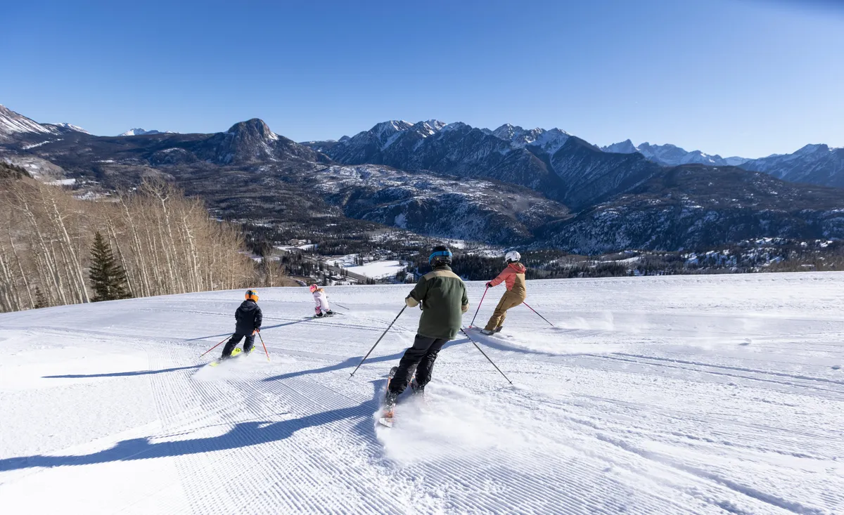 This image captures skiers actively descending a snowy mountain slope beneath a clear blue sky. The snow-covered peaks form a dramatic backdrop, perfectly evoking the thrill and beauty of alpine sports at Purgatory Resort.