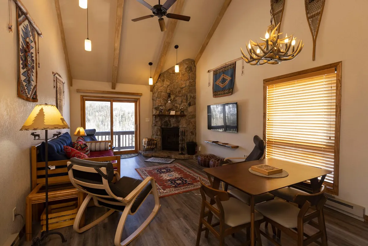The image showcases a cozy condo interior with a striking stone fireplace, a large window offering scenic mountain views, a welcoming dining table, and a classic rocking chair. The warm lighting and inviting furnishings create a comforting ambiance, perfect for relaxing after a day of adventure.