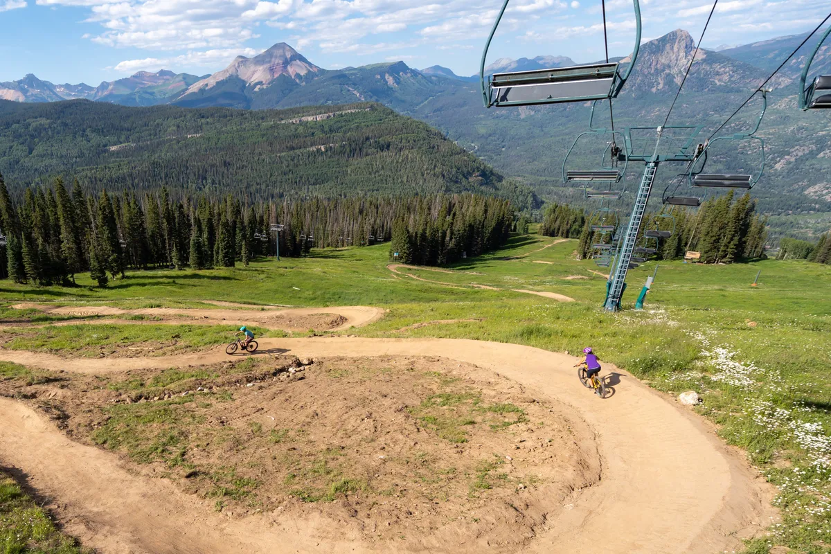 An exterior shot highlighting the dramatic mountain range, with visible bicycle trails and a ski lift, suggesting adventure and exploration right outside the resort. The scene is lively and promises outdoor activities like hiking, biking, and scenic viewing.