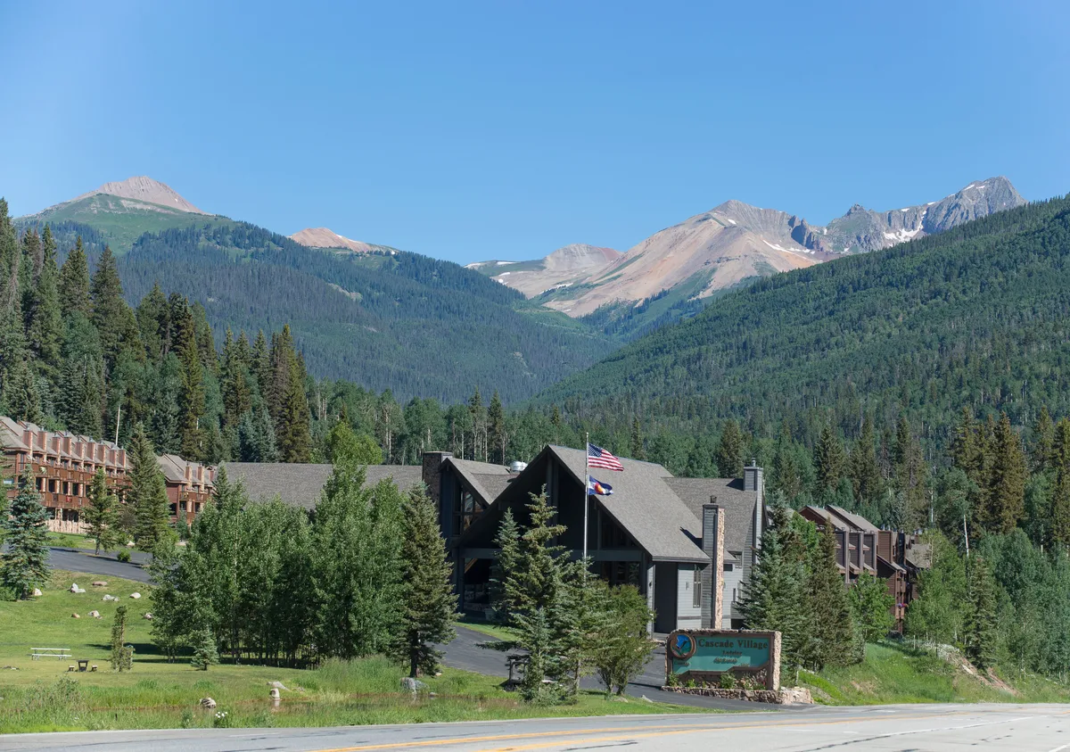 The image shows the main lodge structure nestled against a scenic mountain backdrop, surrounded by trees and inviting landscaping. Flags wave near the entrance, and the overall scene is bustling yet welcoming, hinting at a range of activities and communal experiences.