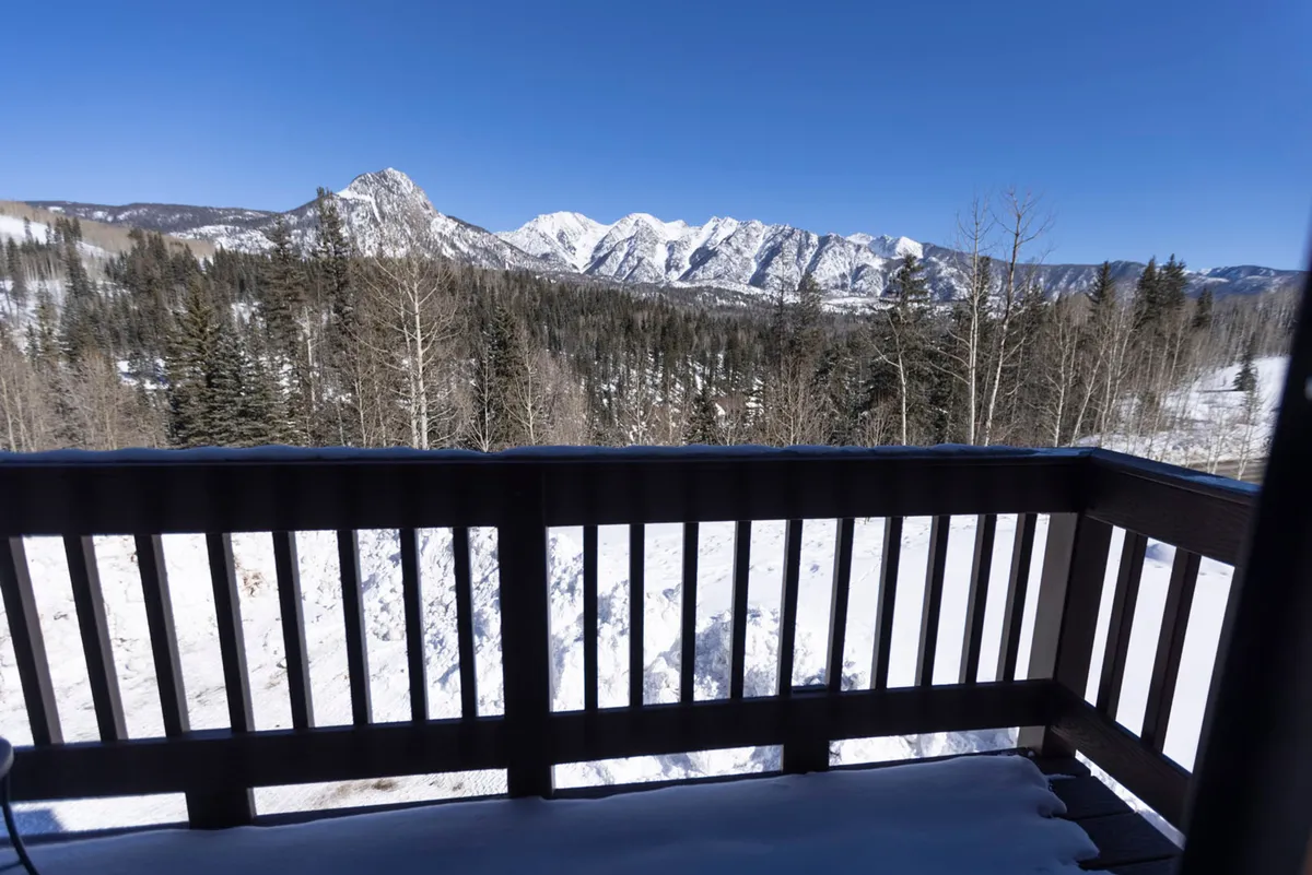 A serene view from a deck or balcony overlooking snow-covered mountains and a dense pine forest. The scene includes a sturdy deck railing and hints that guests can enjoy the outdoors in privacy. The lighting suggests either sunset or early evening, amplifying the romantic and tranquil mood.
