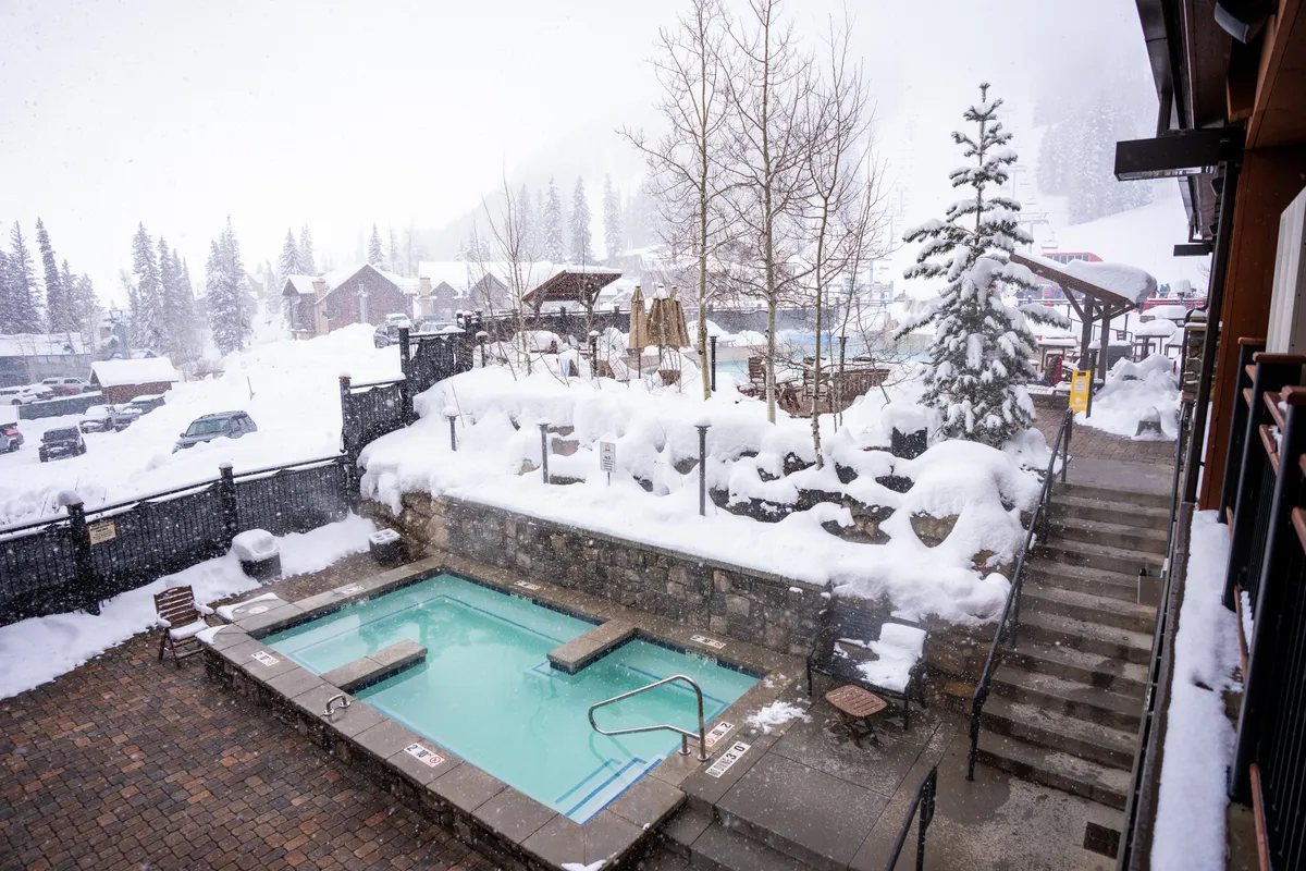 This image showcases the lodge's hot tub surrounded by a beautiful snow-covered landscape and trees. Steps lead up to the steaming water, where guests can relax and socialize while taking in the tranquil, wintry scenery.