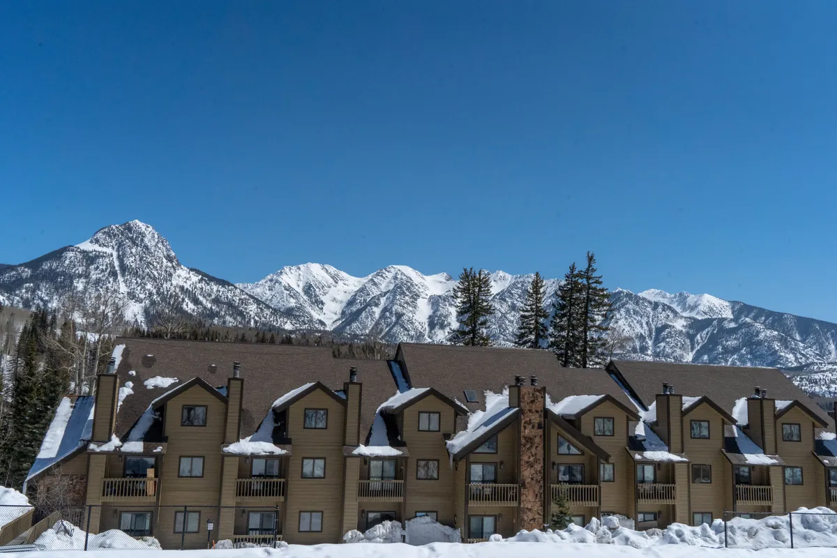 A tranquil exterior scene featuring snow-covered roofs and wooden balconies against a sweeping mountain range. The cozy architecture and stone accents suggest comfort and warmth after adventurous days, while the mountainous backdrop hints at the ski runs nearby.